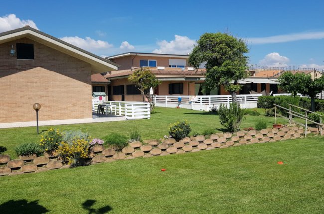 Edificio con giardino ben curato, siepi e fiori sotto un cielo blu.