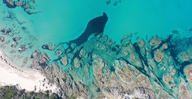 Spiaggia con mare turchese e rocce sommerse viste dall'alto.