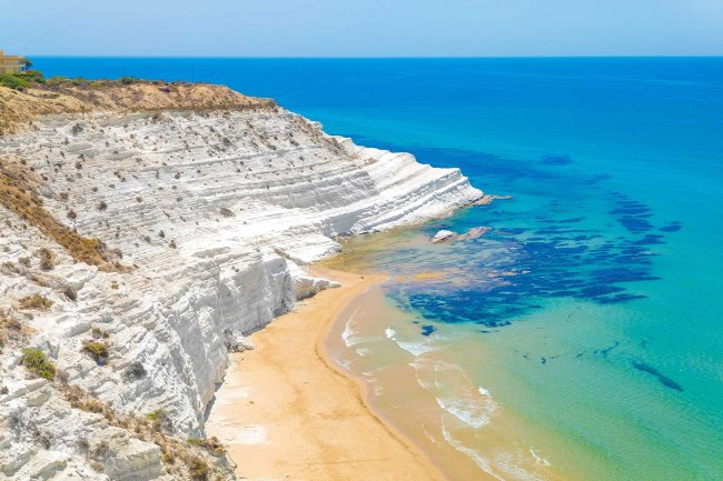 Scogliera bianca su mare turchese e spiaggia dorata.