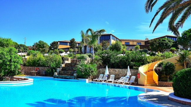 Piscina azzurra con vegetazione lussureggiante e edifici sullo sfondo.