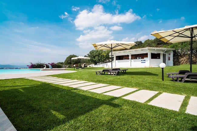 Prato verde, ombrelloni, bar e piscina sullo sfondo.