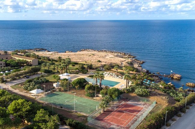 Vista aerea di un resort con campi da tennis e mare.