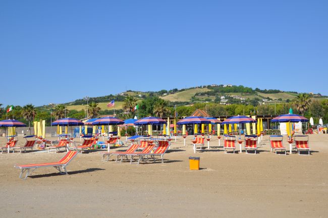 Lettini e ombrelloni colorati su una spiaggia sabbiosa, collina verde sullo sfondo.