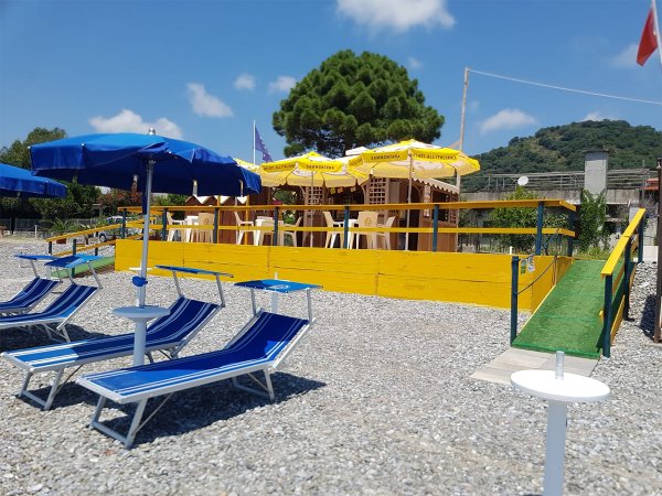 Spiaggia con ombrelloni blu e sdraio di fronte a un chiosco con sedie e tavoli.