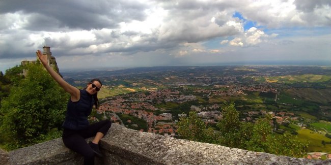 Persona sorridente seduta su un muretto con vista panoramica.