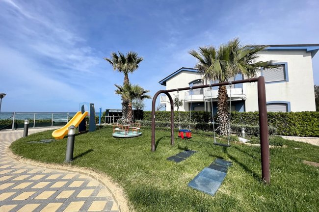 Area giochi con altalene e scivolo vicino a palme.