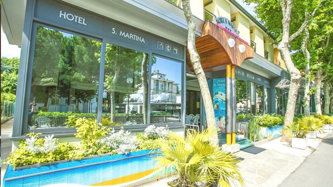 Facciata hotel con ampie vetrate, piante verdi.