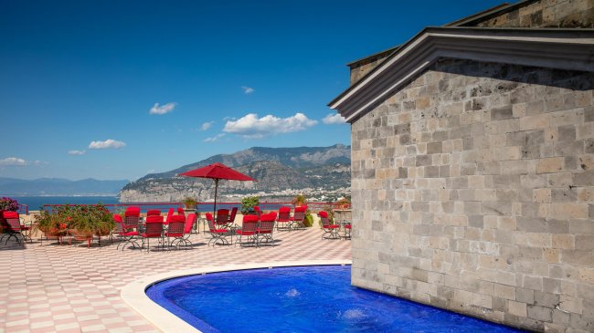 Terrazza con piscina e sedie rosse sotto un cielo blu.