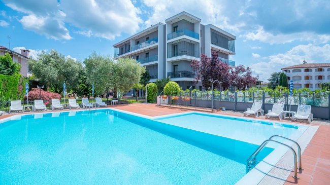 Piscina con hotel moderno sullo sfondo, circondata da lettini.