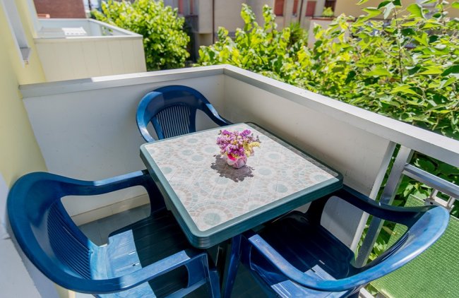 Balcone con tavolino, tre sedie blu e piccoli fiori.