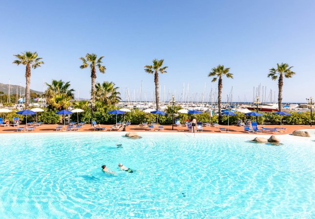 Piscina con due persone che nuotano, palme e barche sullo sfondo.