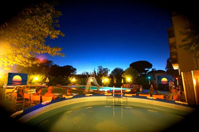 Piscina illuminata di notte con sedie e ombrelloni.