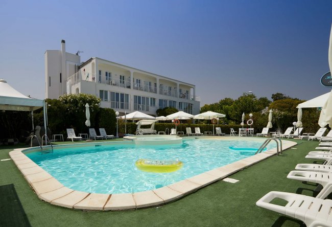 Piscina all'aperto con sdraio bianche e hotel sullo sfondo.