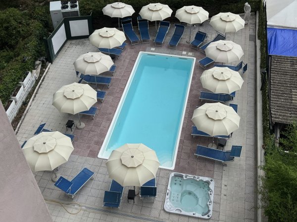 Piscina rettangolare con lettini blu e ombrelloni bianchi intorno.