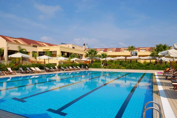 Piscina all'aperto con ombrelloni bianchi e lettini, edificio giallo sullo sfondo.