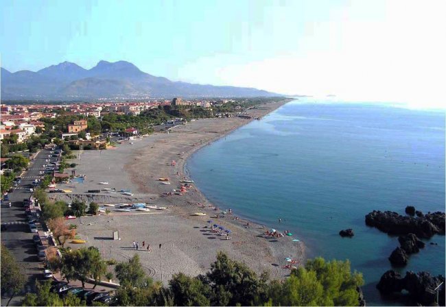 Spiaggia lunga, mare calmo, case e montagne sullo sfondo.