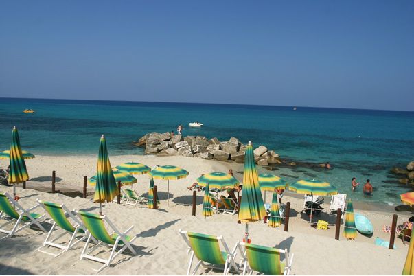 Spiaggia con sedie a sdraio e ombrelloni verdi, rivolti verso un mare azzurro brillante.