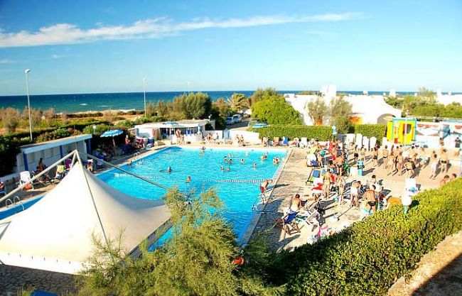 Piscina all'aperto vicino al mare con persona che nuotano e sdraio occupate.