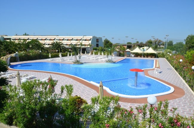 Ampia piscina con fontane e ombrelloni, edificio sullo sfondo.