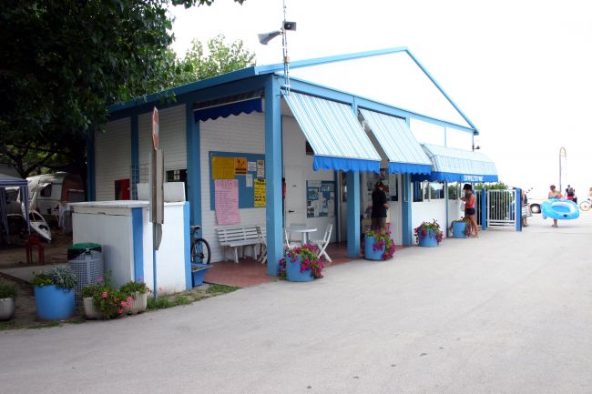 Edificio bianco e blu con tende esterne, piante in vasi azzurri e persone.