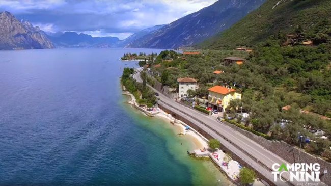 Strada costiera accanto a un lago con spiaggia e montagne in lontananza.