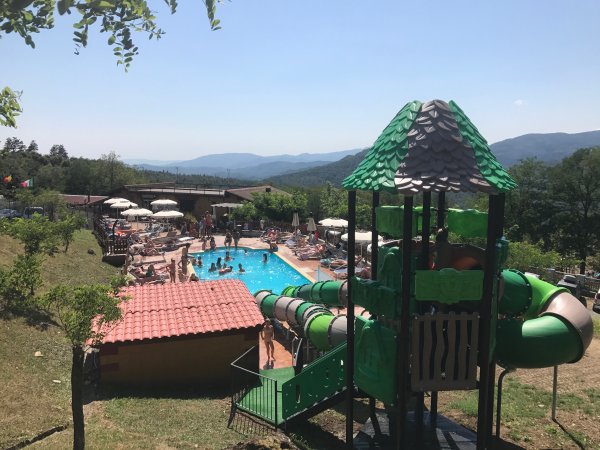 Piscina affollata e area giochi con scivoli, colline sullo sfondo.