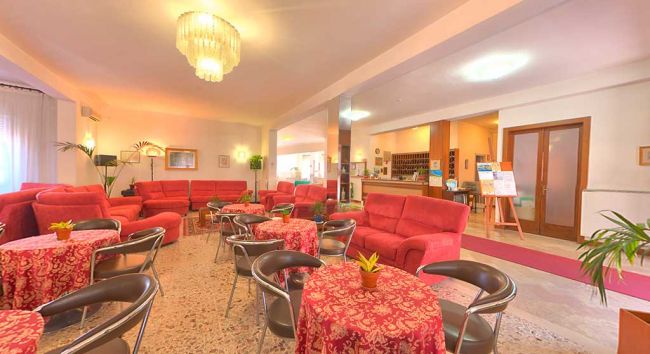 Hall di un hotel con divani rossi, tavolini e un grande lampadario.