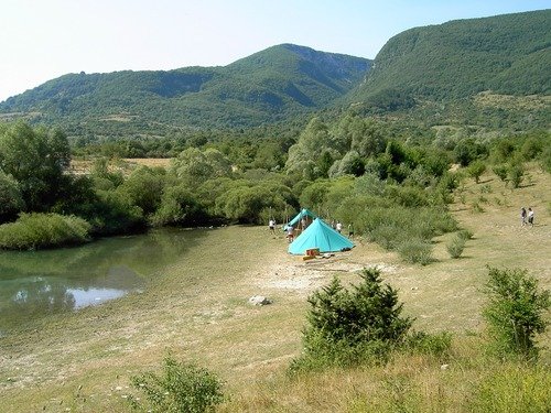 Tenda verde vicino a un piccolo lago, circondata da colline verdi.