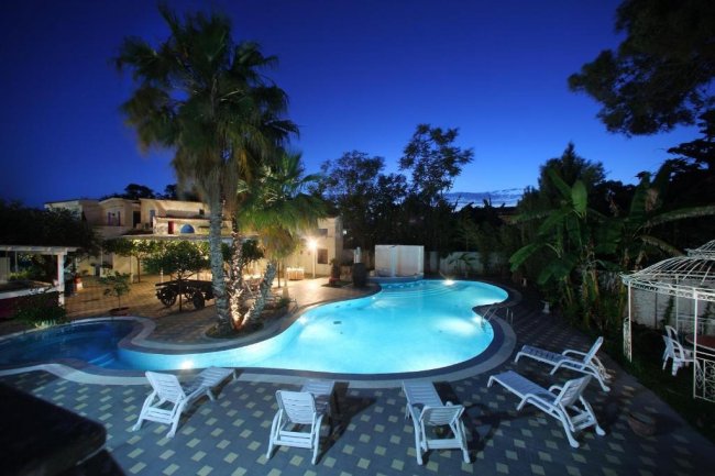 Piscina illuminata di notte con palme e sedie a sdraio.