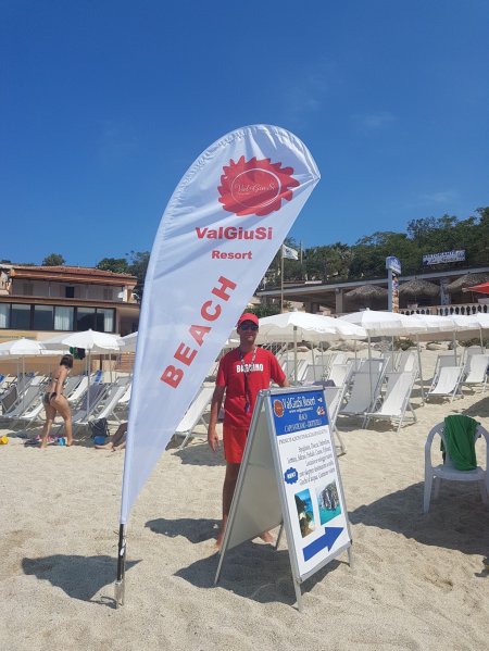 Spiaggia con lettini bianchi e un addetto davanti a una bandiera.