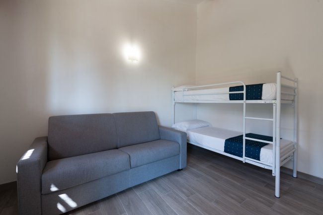 Stanza con divano grigio e letto a castello bianco, illuminata da una luce soffusa.