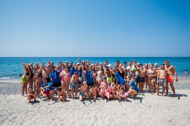 Gruppo di persone sorridenti sulla spiaggia, con il mare azzurro sullo sfondo.