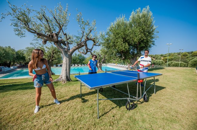 Due persone giocano a ping pong all'aperto mentre una ragazza osserva.