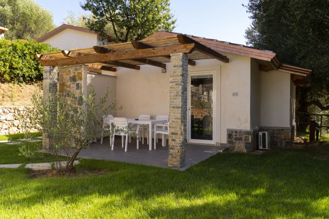 Un bungalow con veranda, tavolo bianco e sedie, circondato da prato verde e alberi.