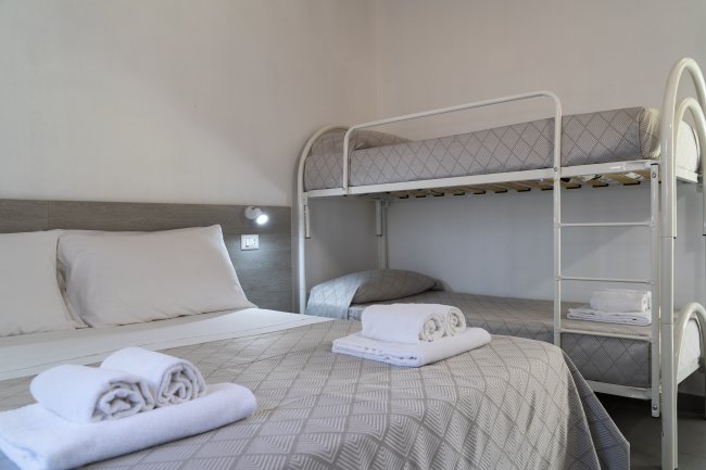 Dettaglio di un letto matrimoniale e letti a castello con lenzuola grigie e asciugamani bianchi.