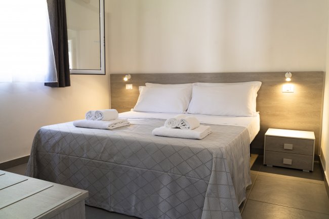 Camera da letto con letto matrimoniale, coperte grigie e due comodini con luci accese.