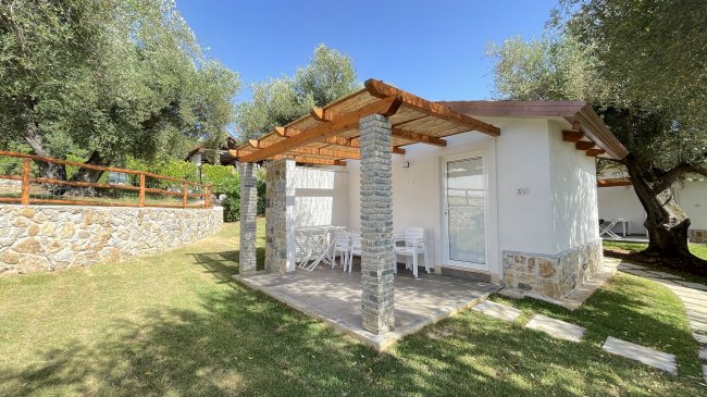 Bungalow bianco con tettoia in legno, veranda con tavolo e sedie. Circondato da erba e alberi.