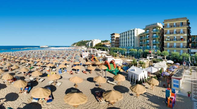 Spiaggia affollata con ombrelloni e hotel sullo sfondo.