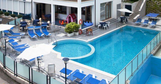 Piscina con lettini blu e tavoli con ombrelloni bianchi.