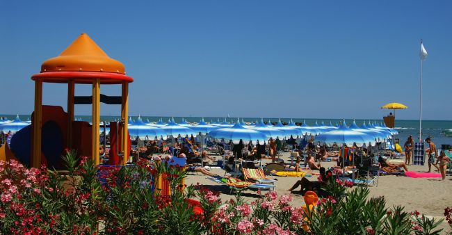 Spiaggia affollata con ombrelloni blu e bianchi, un parco giochi colorato e piante fiorite.