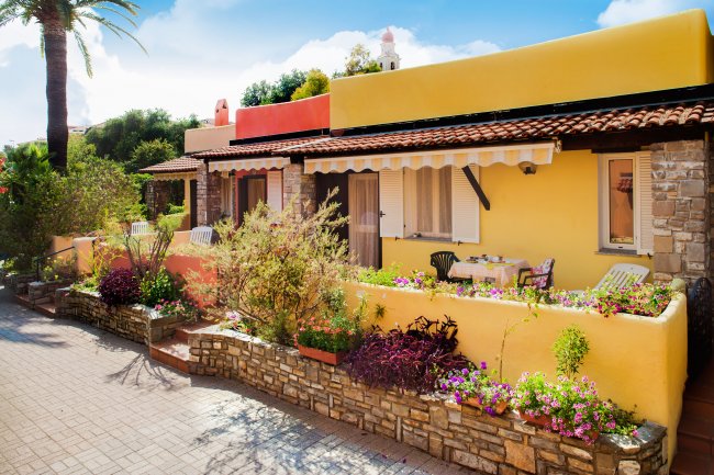 Casetta gialla con tetto in tegole, veranda con fiori colorati e sedie a sdraio.