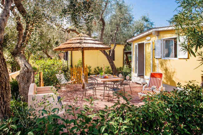 Patio all'aperto con tavolo, sedie, ombrellone di paglia e piante rigogliose attorno.