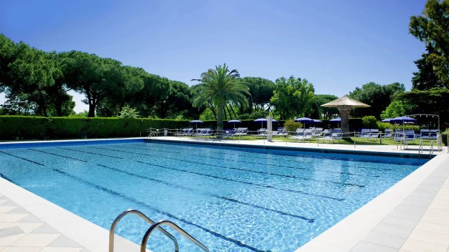 Piscina rettangolare con ombrelloni e alberi sullo sfondo.