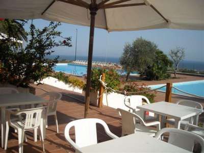 Tavoli e sedie bianche sotto ombrelloni, vista piscina e mare.