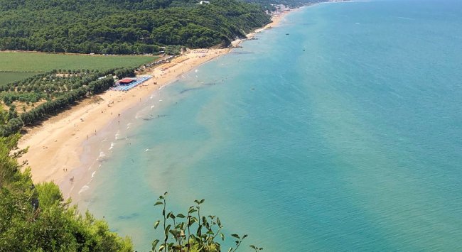 Lunga spiaggia vista dall'alto, con mare turchese e colline verdi sullo sfondo.