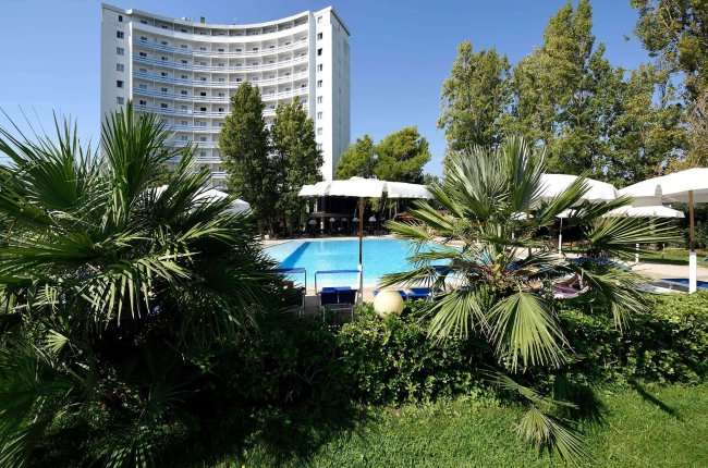Edificio alto con piscina, palme e ombrelloni.