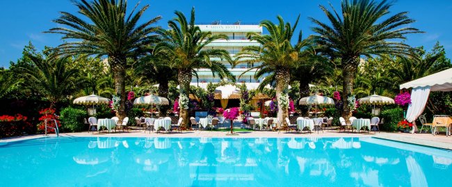 Piscina circondata da palme e tavolini con ombrelloni.