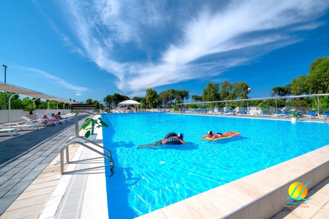 Piscina all'aperto con persone su materassini gonfiabili e cielo parzialmente nuvoloso.
