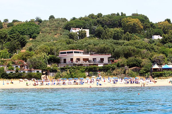 Spiaggia con ombrelloni e sdraio davanti a una casa tra alberi verdi.