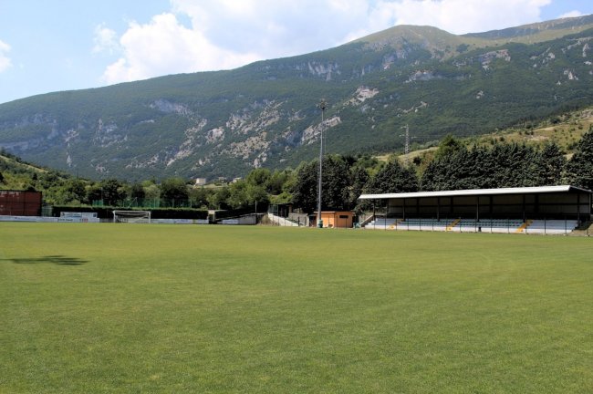 Campo di calcio con tribune e montagne sullo sfondo.